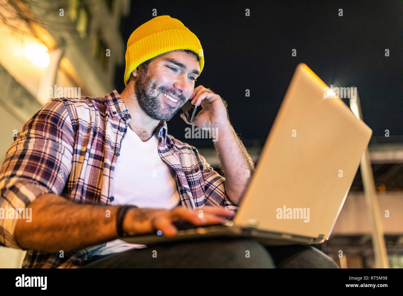 UK, London, smiling man using phone and laptop out in the city at night Stock Photo