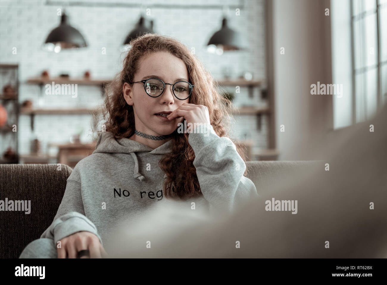 Teenage girl. Curly appealing dark-haired teenage girl wearing grey hoodie sitting on sofa Stock Photo