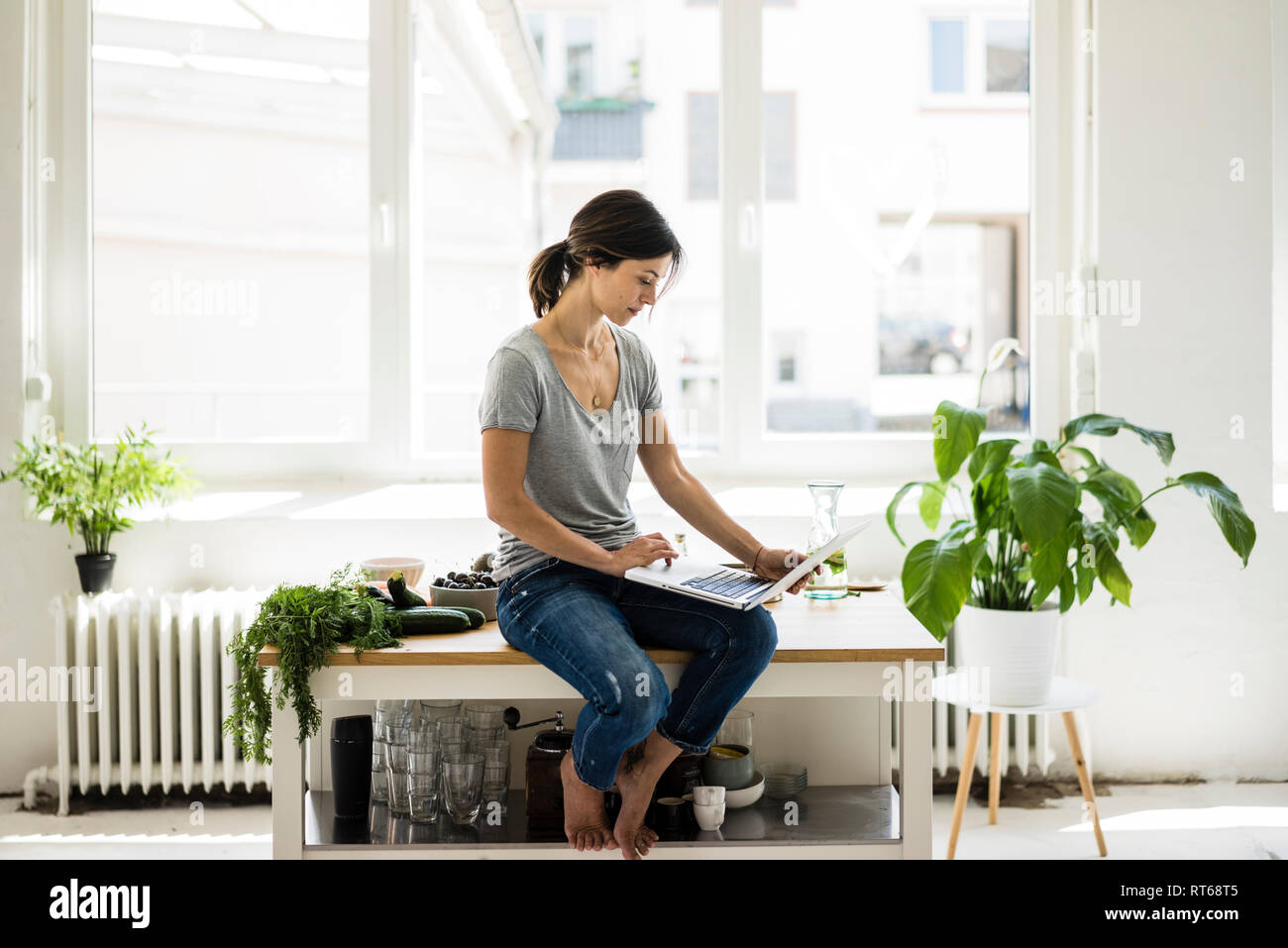 Woman sitting on kitchen table, searching for healthy recipes, using laptop Stock Photo