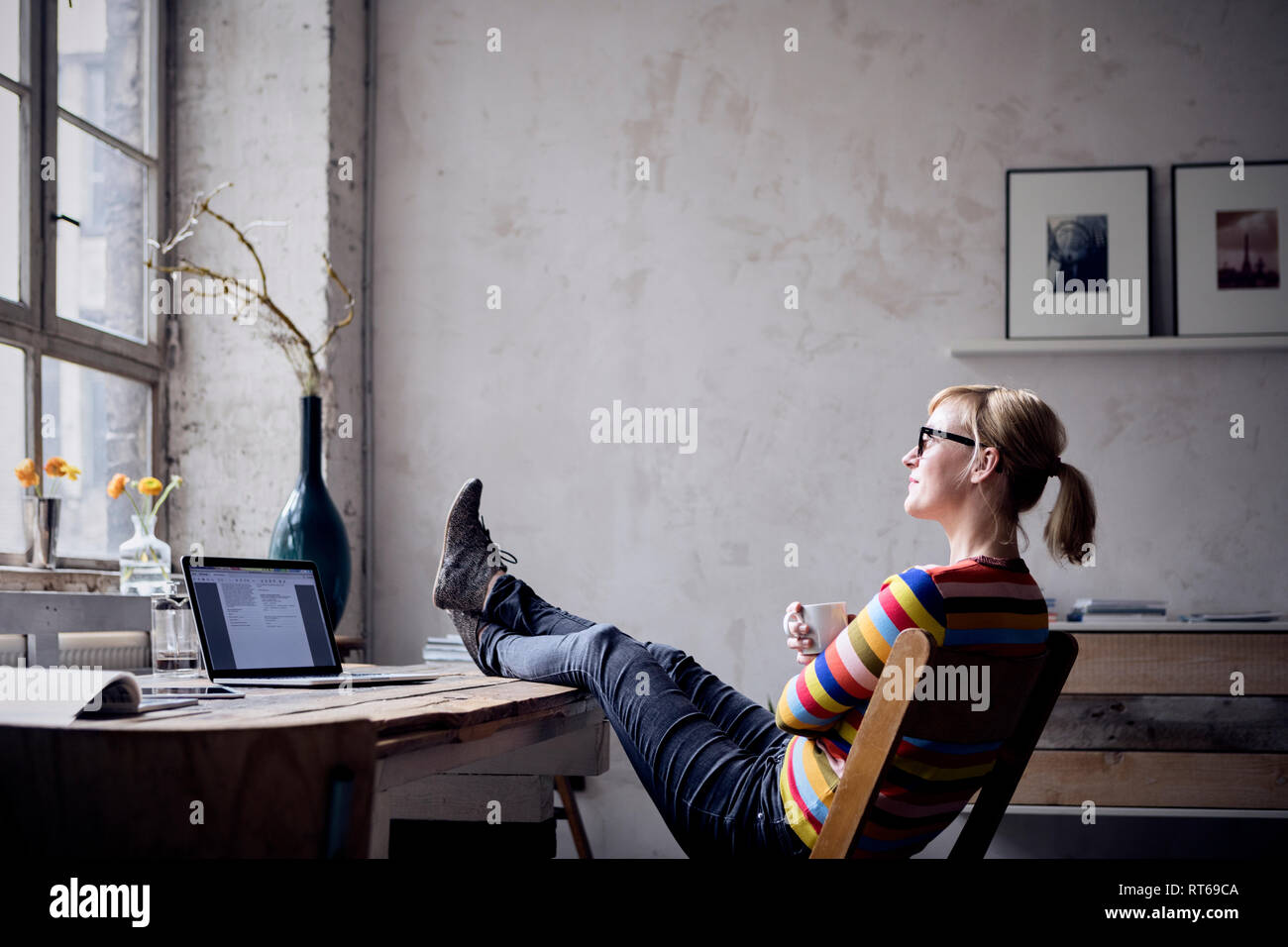 Smiling woman sitting with feet up at desk in a loft looking through window Stock Photo