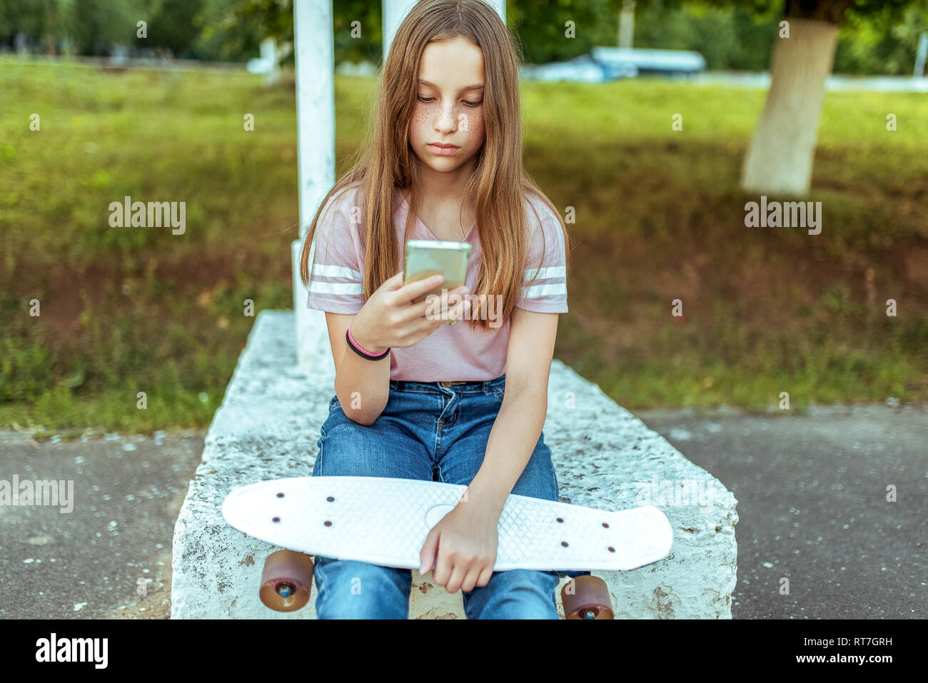 Teen girl 12-13 years old, sitting in hands of a skate. In summer in city in casual jeans and pink T-shirt. Communication in Internet, in hands Stock Photo