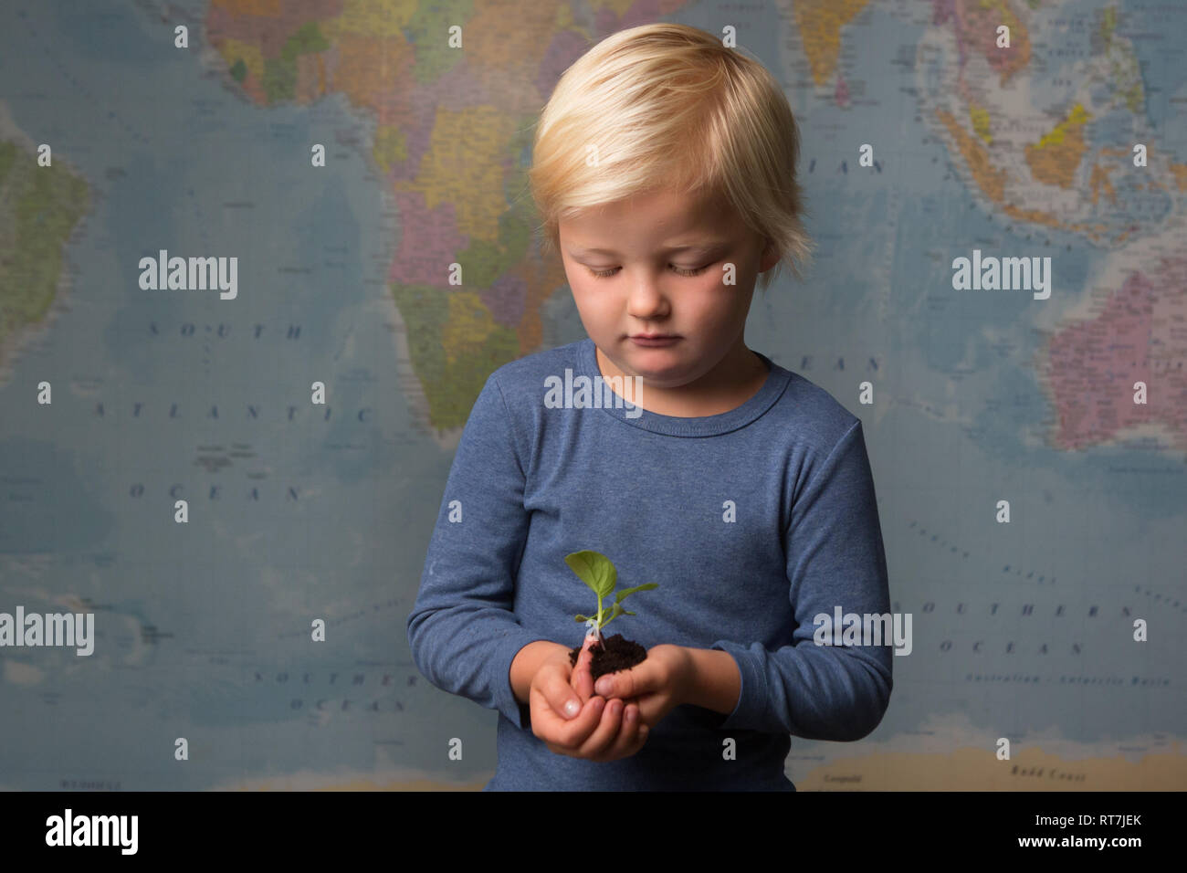 Cute blonde child carefully holds a seedling in front of a world map Stock Photo