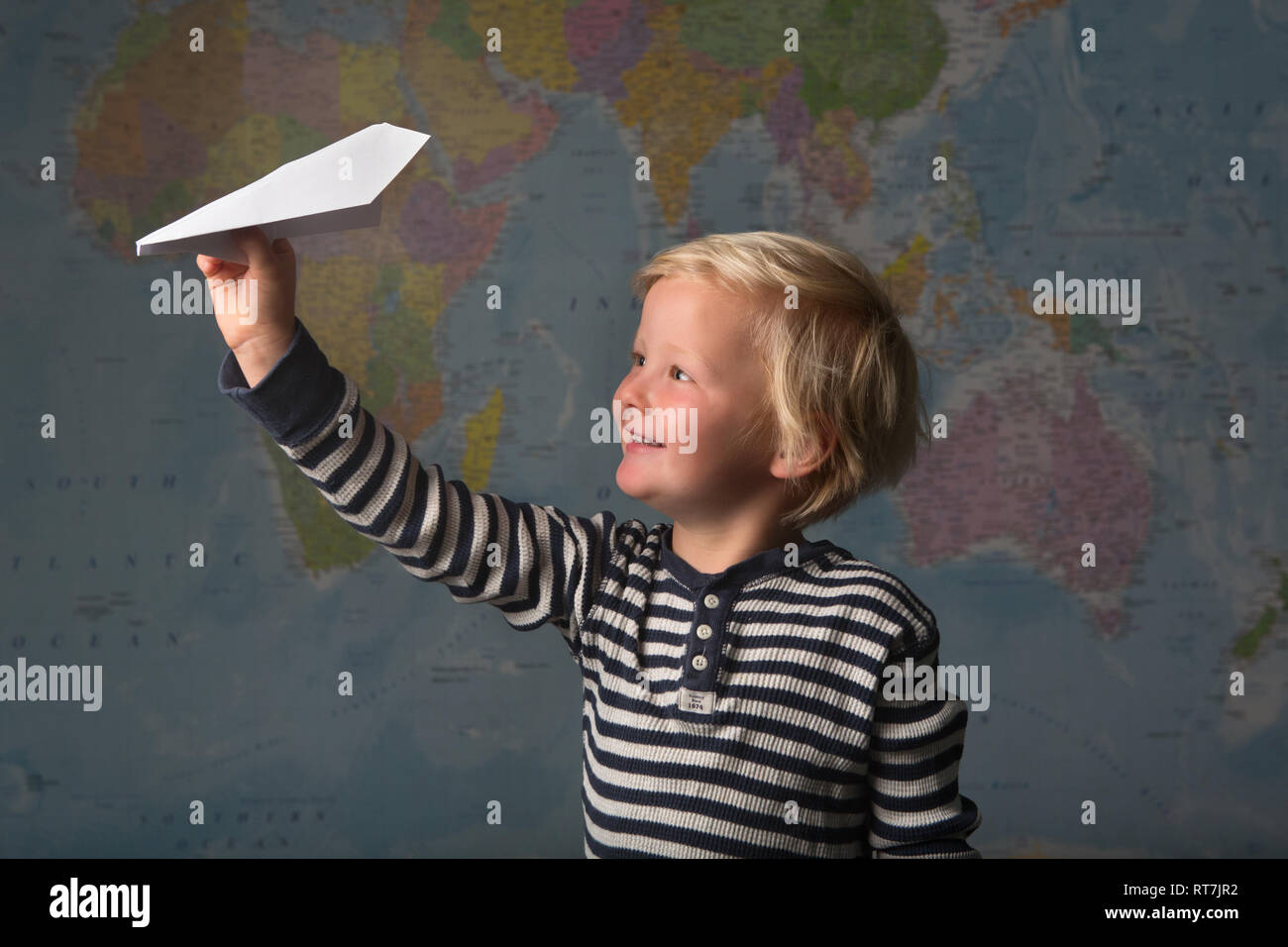 Cute blonde child excitedly flies a paper plane in front of a large world map Stock Photo