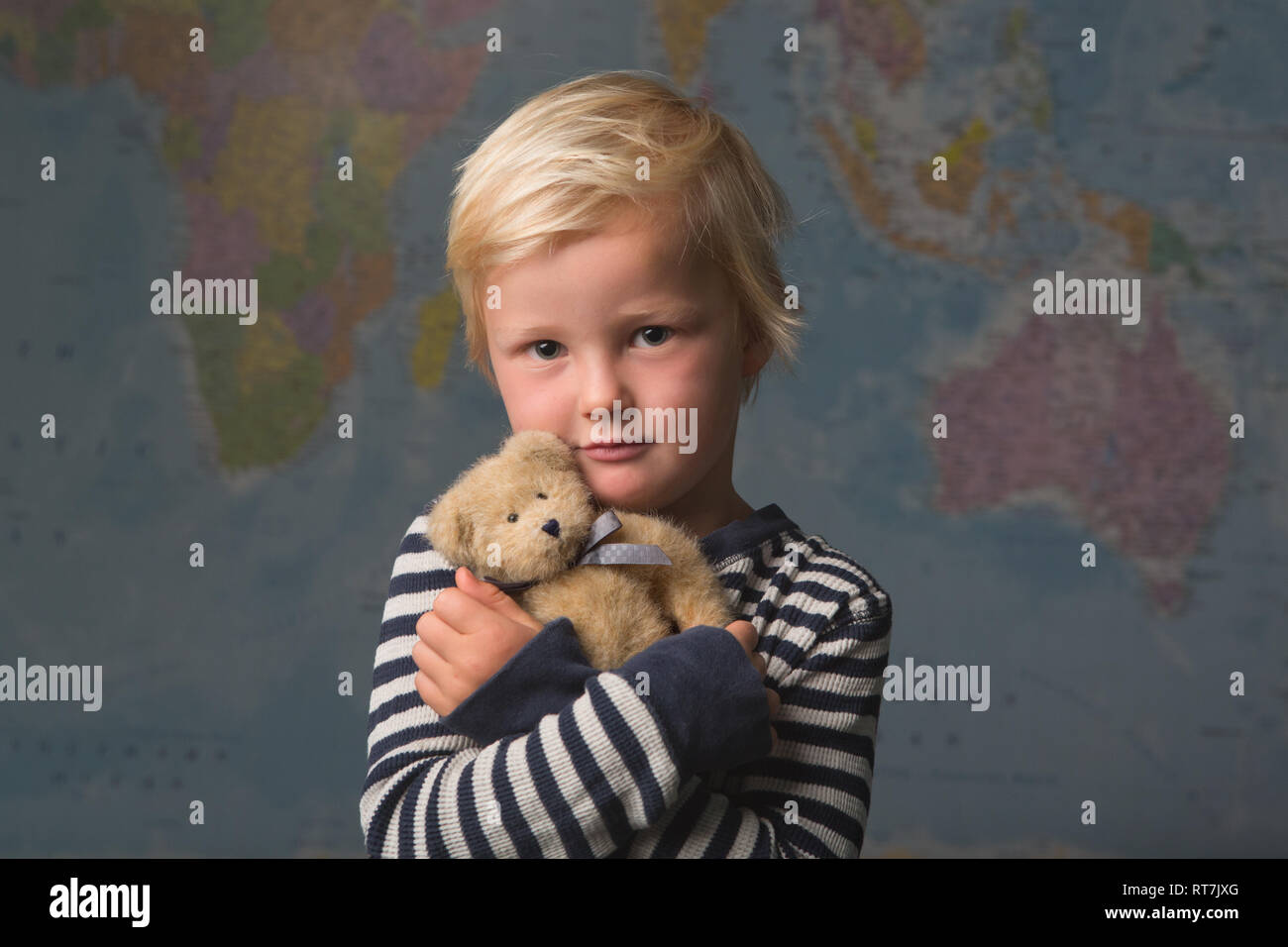 Cute blonde child cuddles his teddy bear in front of a large world map Stock Photo