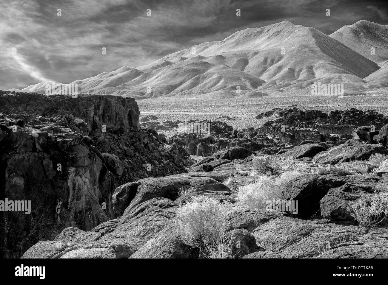 Rocky fields, rock canyon with steep rock walls and barren desert mountains beyond under cloudy skies. Infrared image, black and white. Stock Photo