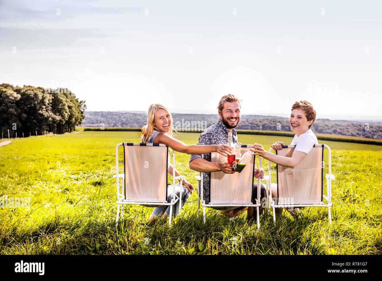 Happy friends sitting on camping chairs in rural landscape clinking bottles Stock Photo