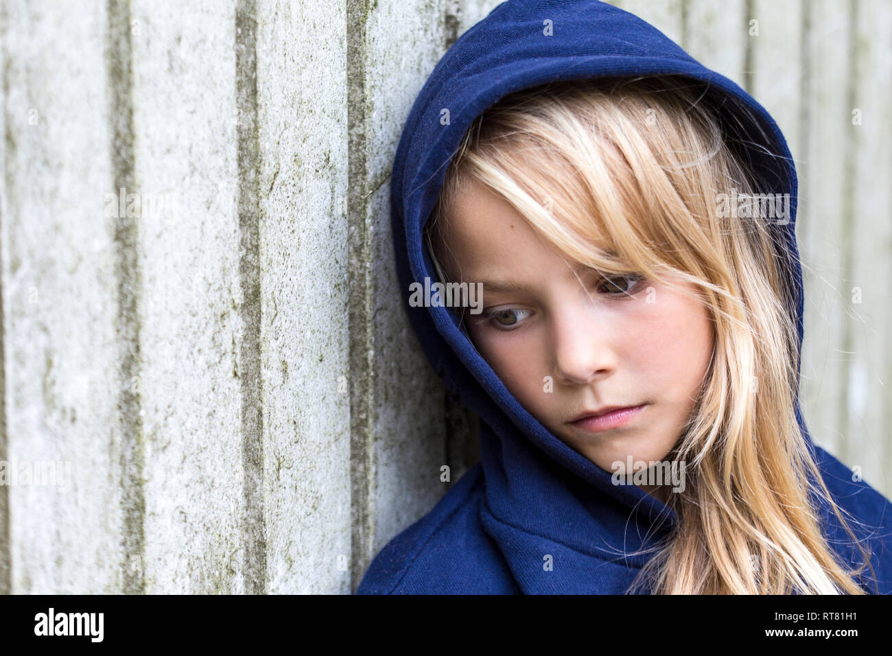 Portrait of sad blond girl wearing blue hooded jacket leaning against wooden wall Stock Photo