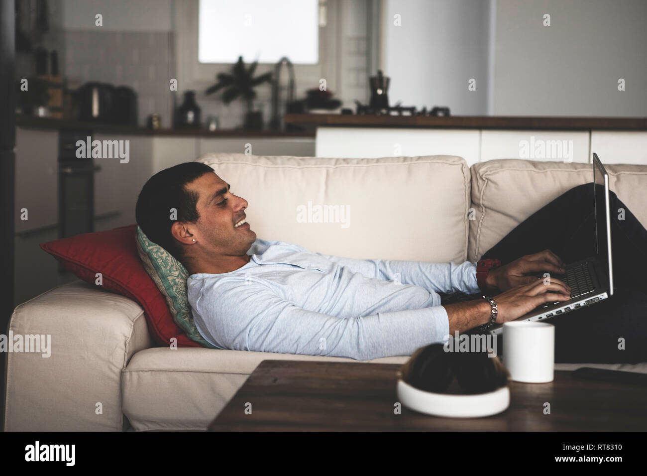 Smiling young man lying on sofa at home using laptop Stock Photo