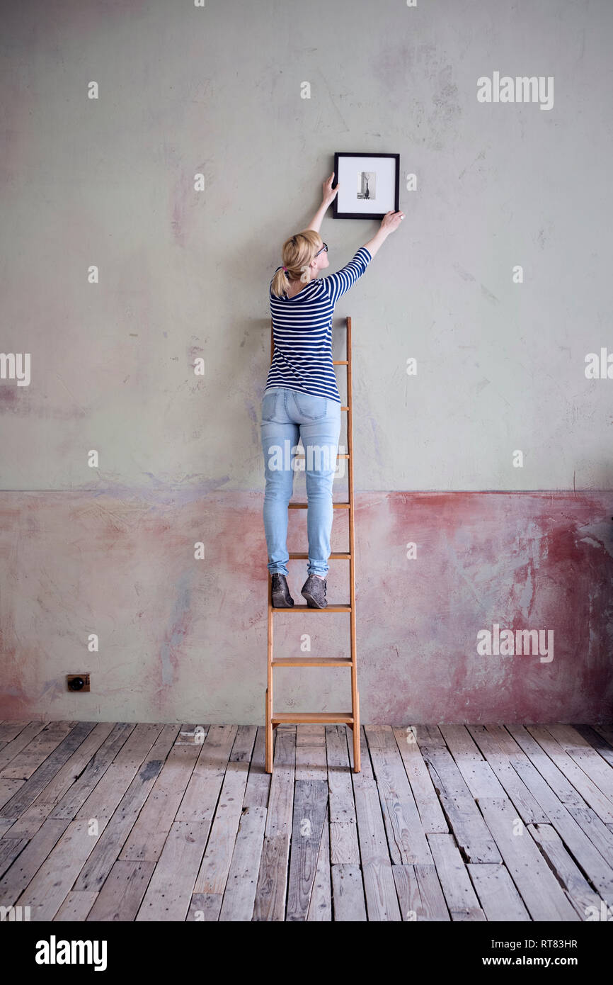 back view of woman on ladder hanging up picture frame in an unrenovated room of a loft Stock Photo