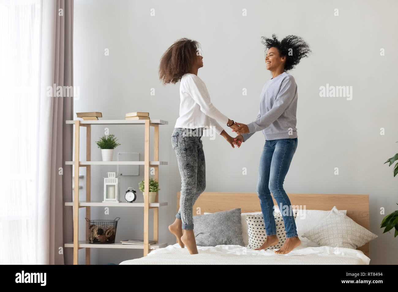Black mother and adolescent daughter holding hands jumping on bed Stock Photo
