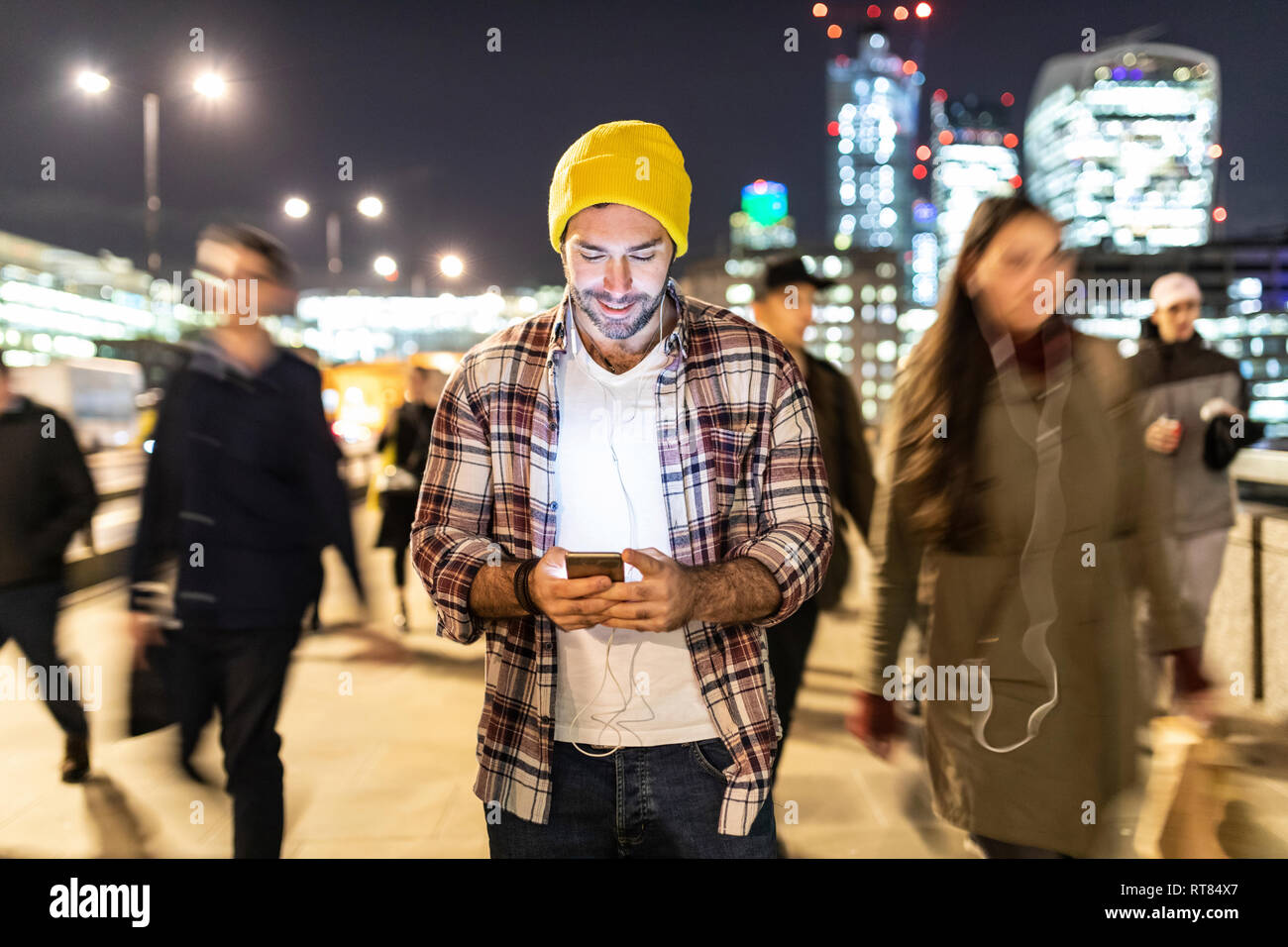UK, London, smiling man looking at his phone by night with blurred people passing nearby Stock Photo
