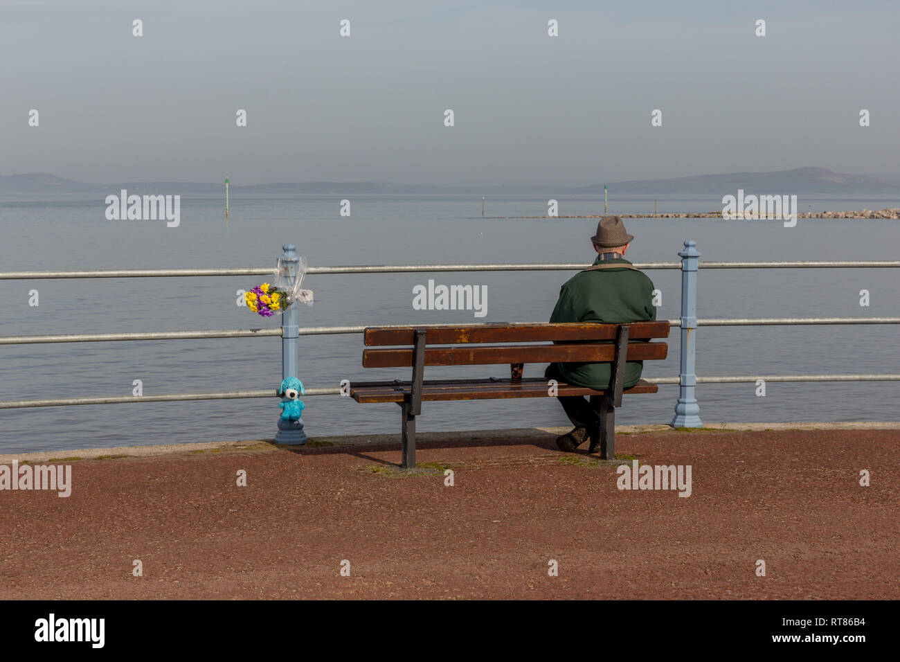 A lone man sat on a promenade bench looking out across Morecambe Bay next to a floral tribute Stock Photo