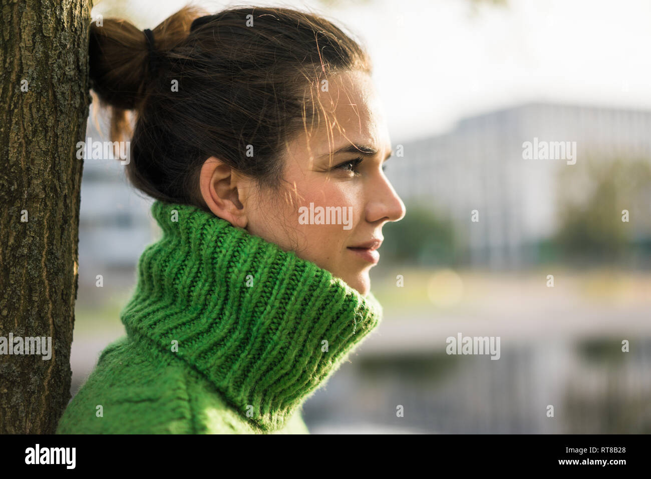 Profile of relaxed woman wearing green turtleneck pullover leaning against tree trunk Stock Photo