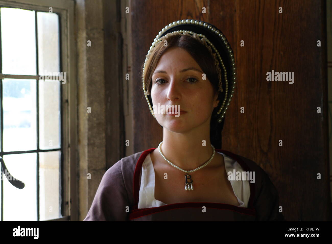 A beautiful young Woman dressed as Anne Boleyn sits by a window at the Tower of London and looks thoughtful. Stock Photo