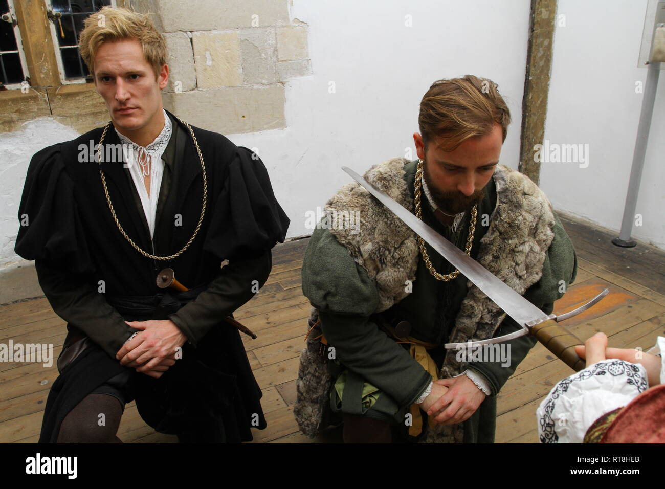 Two young men dressed in authentic Tudor clothing Kneel in front of a sword to be knighted- they wear regal clothes and have a sword on their neck Stock Photo