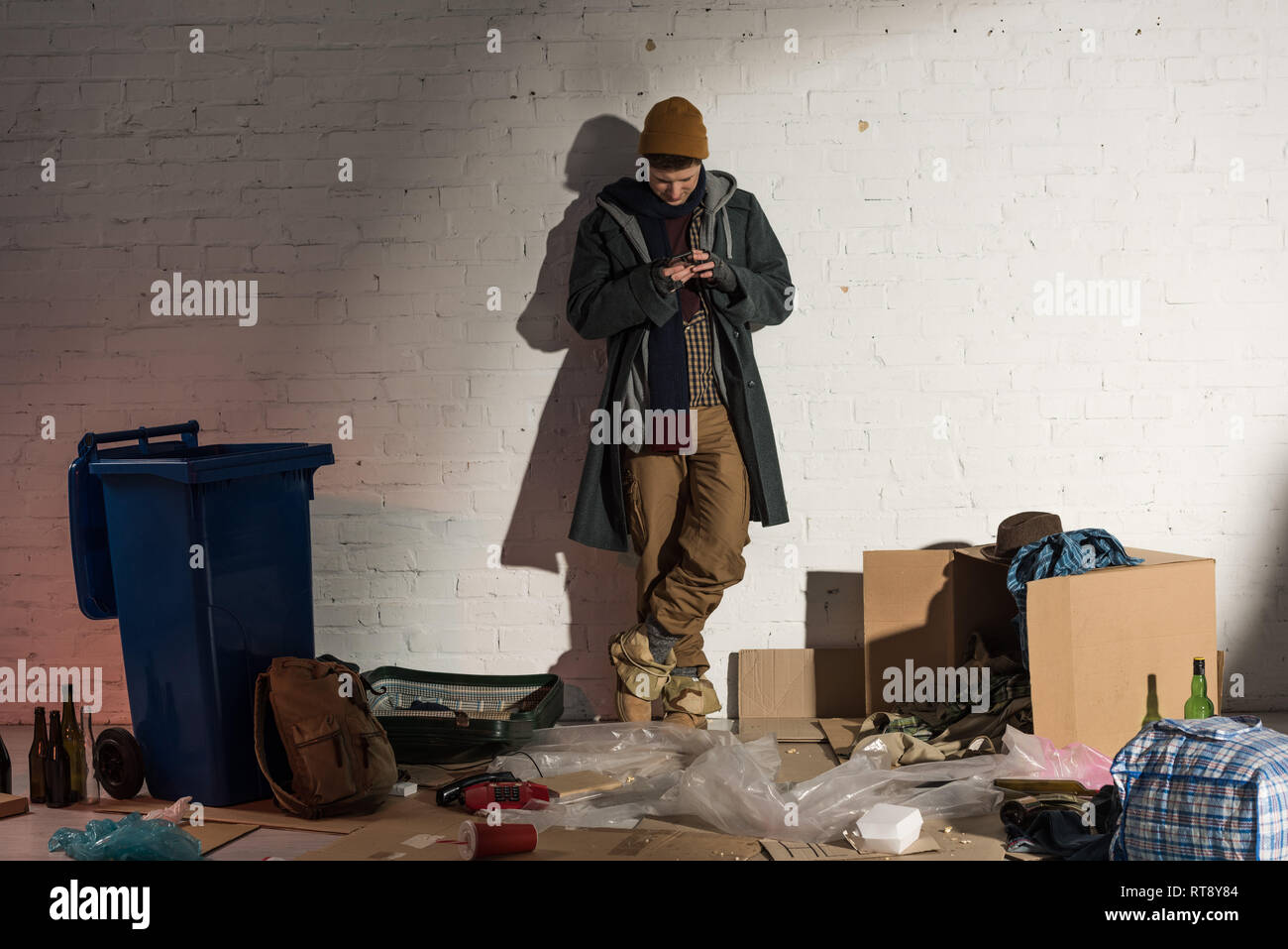 homeless man using smartphone while standing by white brick wall ...