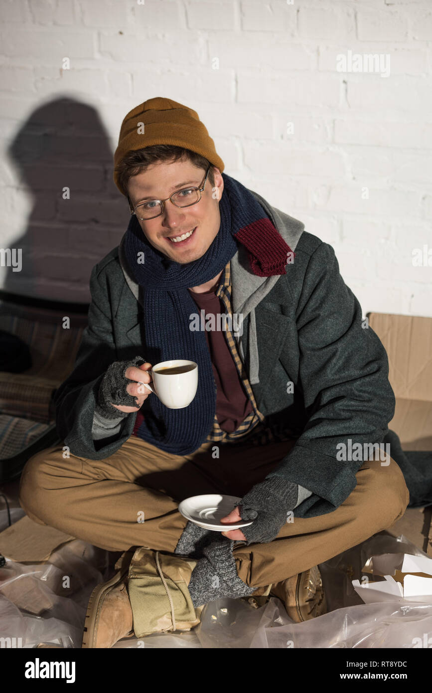 smiling homeless man drinking coffee while sitting by white brick wall