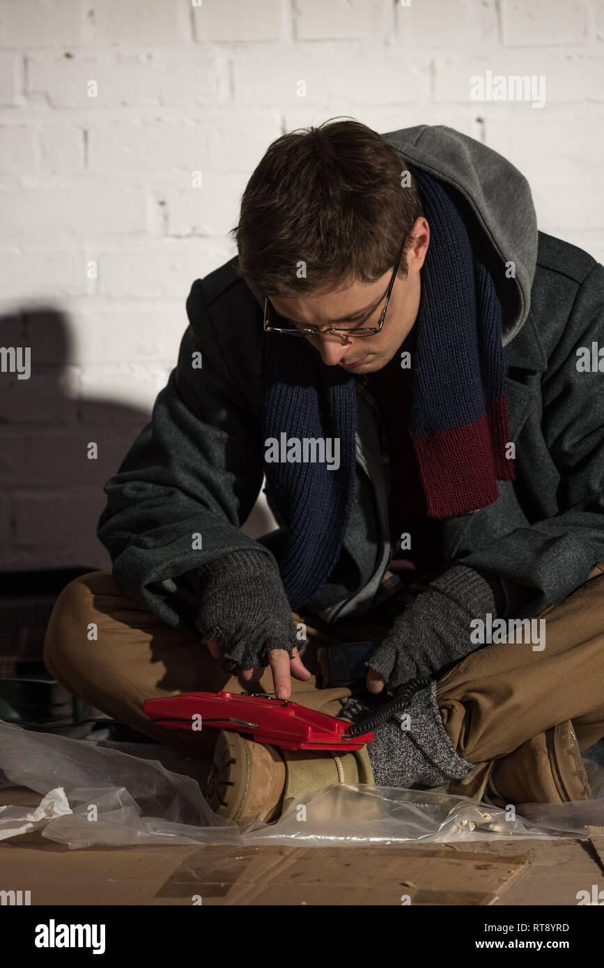 homeless man in glasses using vintage phone while sitting by brick wall ...