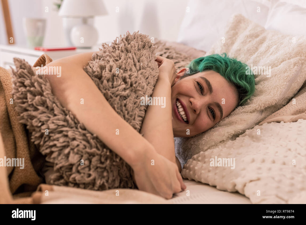 Cheerful woman hugging little fluffy pillow while chilling in bed Stock Photo