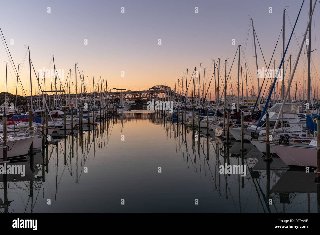 A sunset shot of a marina with the Auckland Harbour bridge on the background, looking down a row of sailing boats Stock Photo