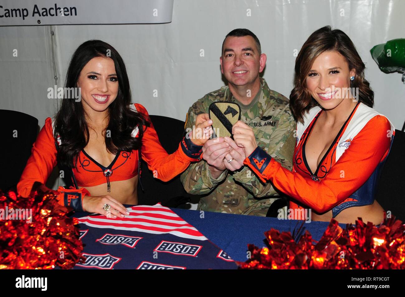 U.S. Army Chief Warrant Officer 3 Michael Hamilton assigned to the 1st Armored Brigade Combat Team, 1st Cavalry Division poses with cheerleaders holding his 1st Cavalry patch as part of a USO meet and greet with the Denver Broncos cheerleaders, mascot and Alumni football players at Camp Aachen's USO annex in Grafenwoehr, Germany, Jan. 26, 2019.   IRONHORSE deployed in support of Atlantic Resolve, an enduring exercise to improve interoperability between U.S. Forces, their NATO allies and partners. Stock Photo