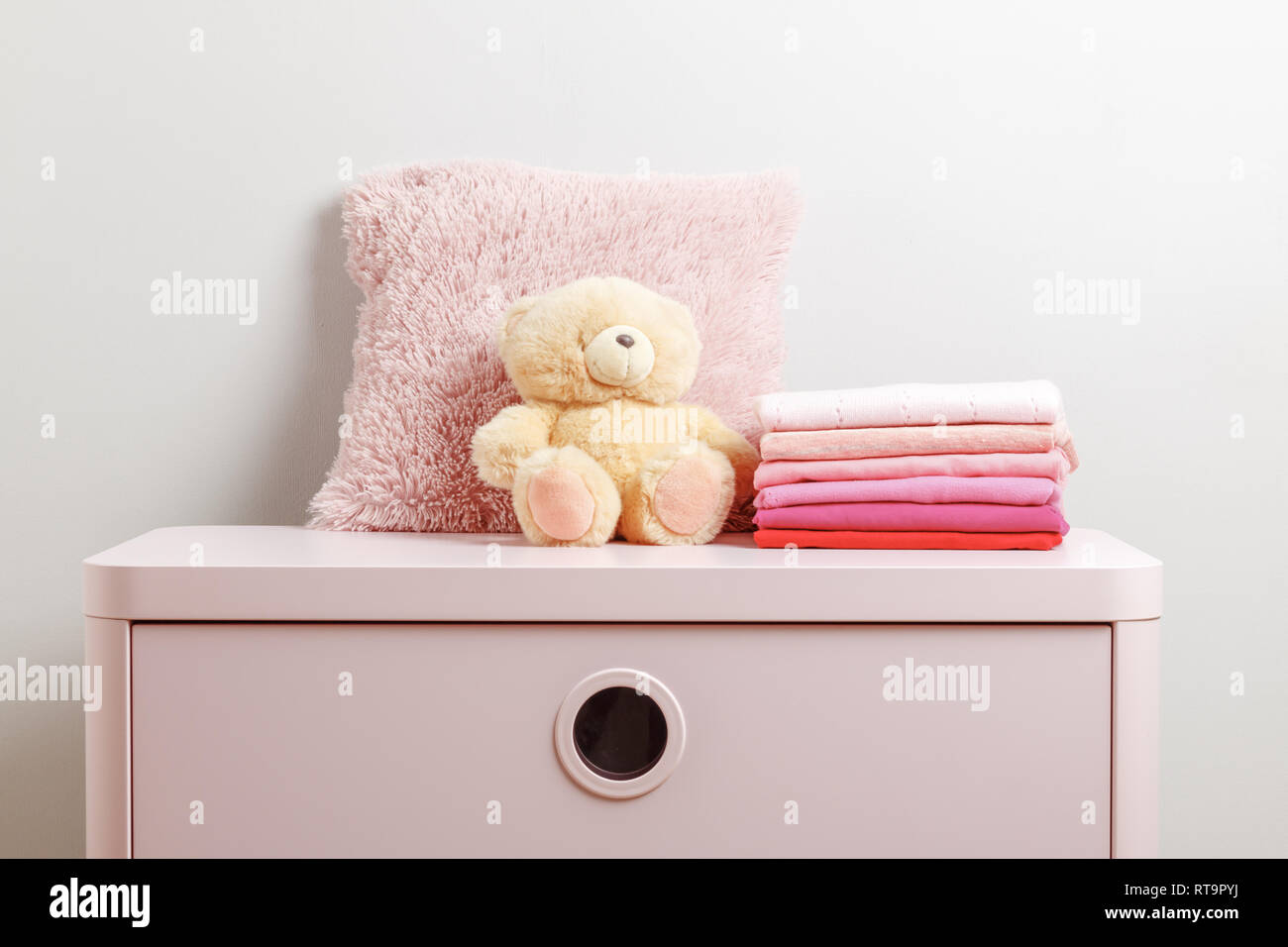 A Stack Of Baby Clothes And A Teddy Bear On A Pink Dresser Gray