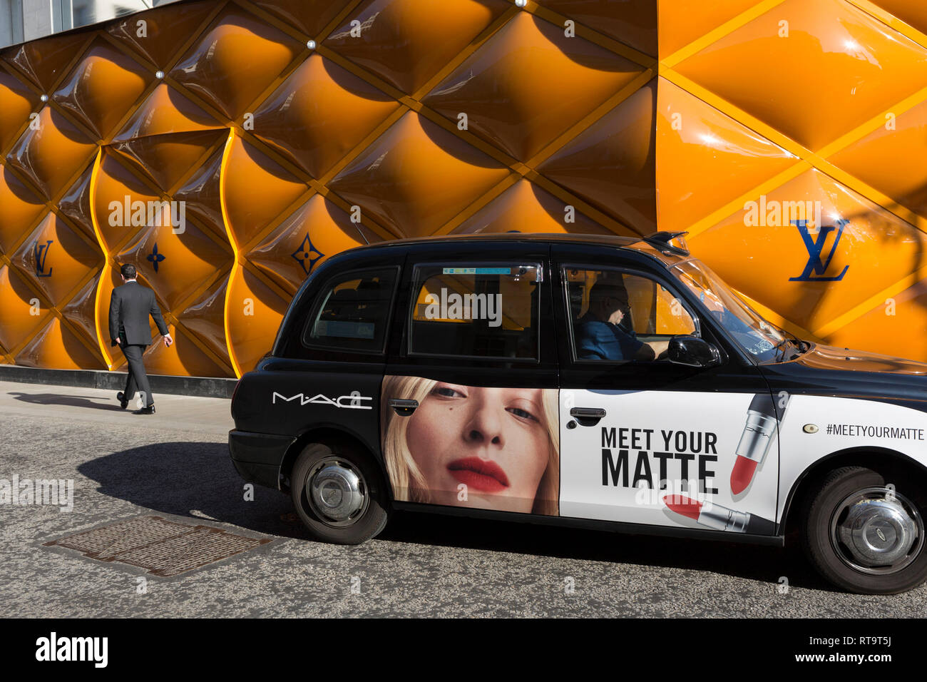 A black cab with advertising for a lipstick brand on the door drives past  the temporary renovation hoarding of luxury brand Louis Vuitton in New Bond  Street, on 27th February 2019, in