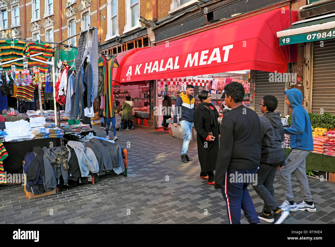 People walk past  AK Halal Meat butcher store & clothing stall, Brixton street market Electric Avenue Brixton South London SW9 England UK KATHY DEWITT Stock Photo