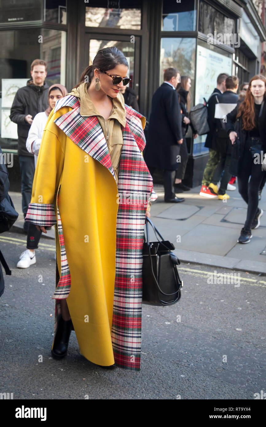 LONDON - FEBRUARY 15, 2019: Stylish attendees gathering outside 180 The Strand for London Fashion Week. Girl in a yellow long coat and a plaid scarf p Stock Photo