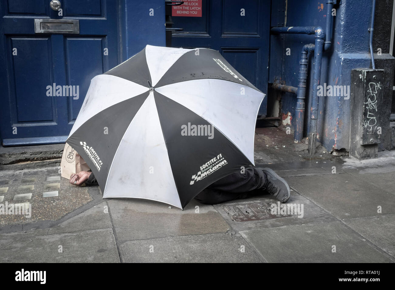 Homeless man using large umbrella for shelter on central London street, London, UK Stock Photo