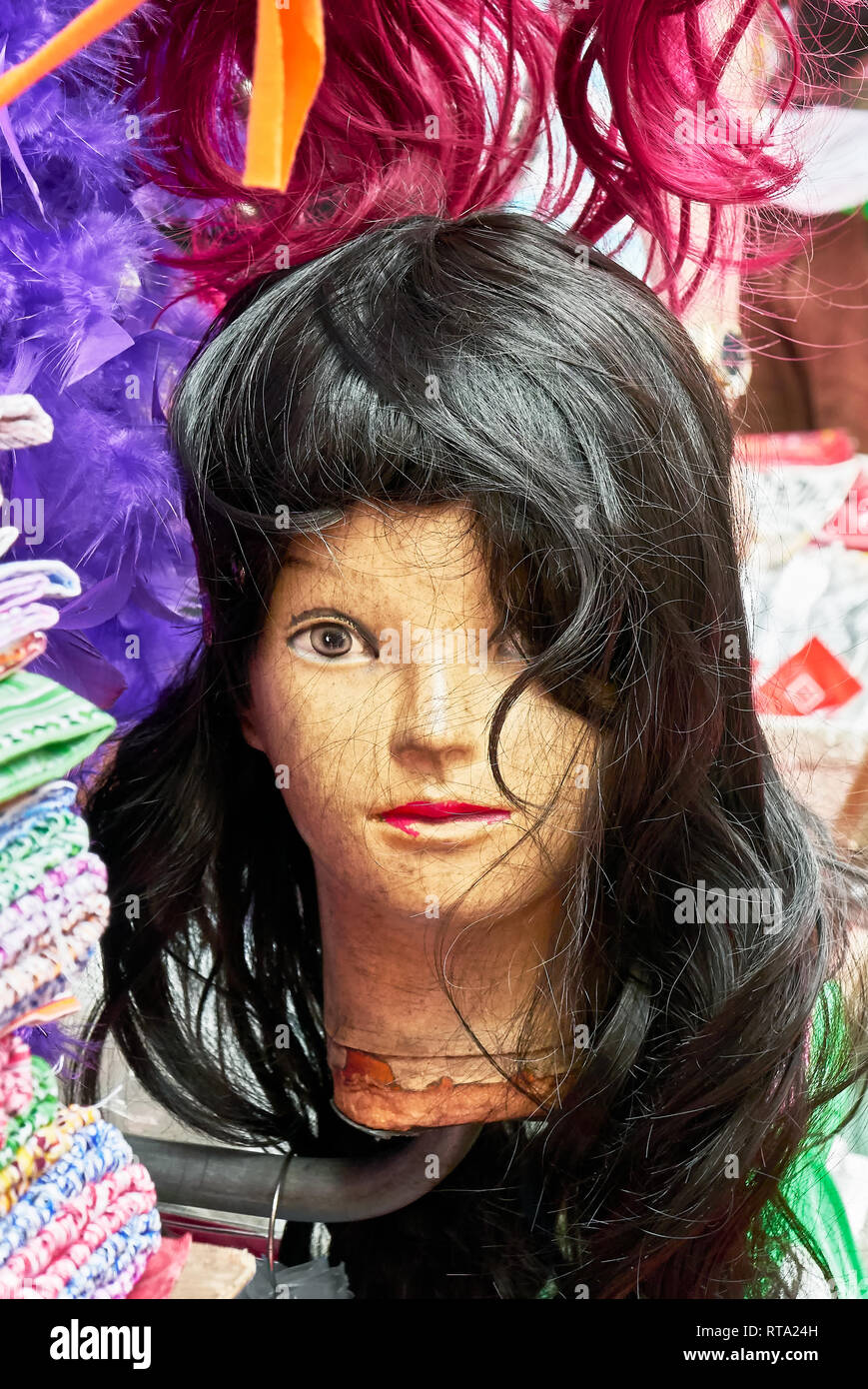 One female young looking mannequin head, looking straight into the camera, is wearing a black long wig, surrounded by red colored hair and clothes Stock Photo