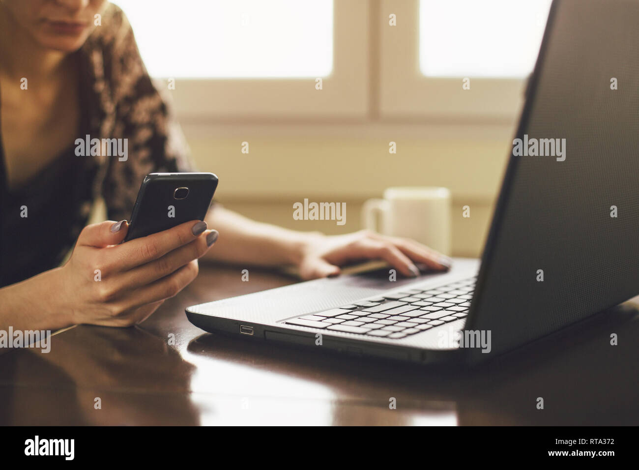 Unrecognizable young woman looking at smartphone while using laptop Stock Photo