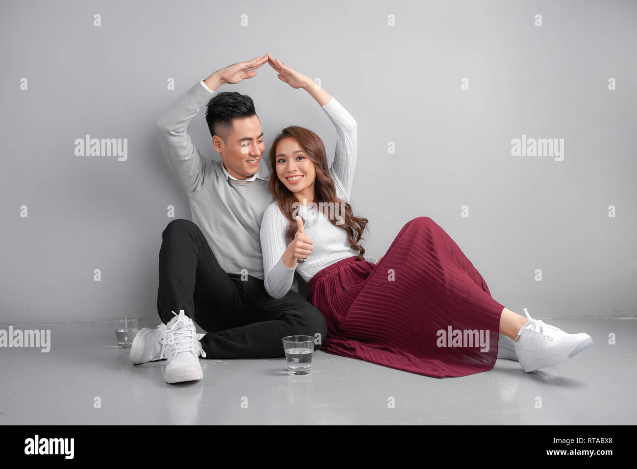 New building residential house purchase apartment concept. Stylish young family sitting on floor, wife and husband making roof figure with hands arms  Stock Photo