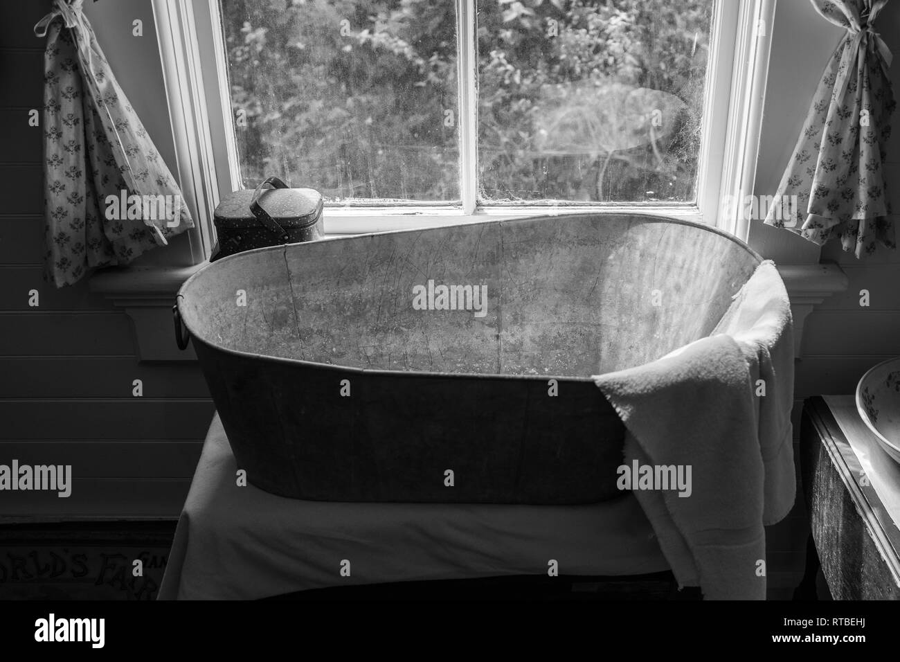 An old school metal bath tub in the window of a house, with a towel draped over the end of the tub, shot in black and white to give a true vintage fee Stock Photo