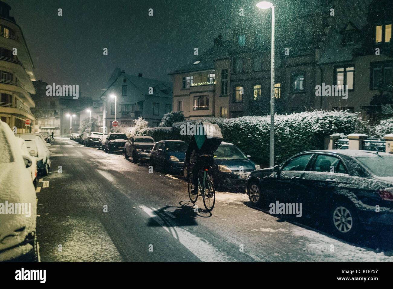 STRASBOURG, FRANCE - DEC 3 2017: Deliveroo delivery employee on bike in French city cycling fast for food delivery on time on a cold winter snowy night in residential neighborhood with cars parked Stock Photo