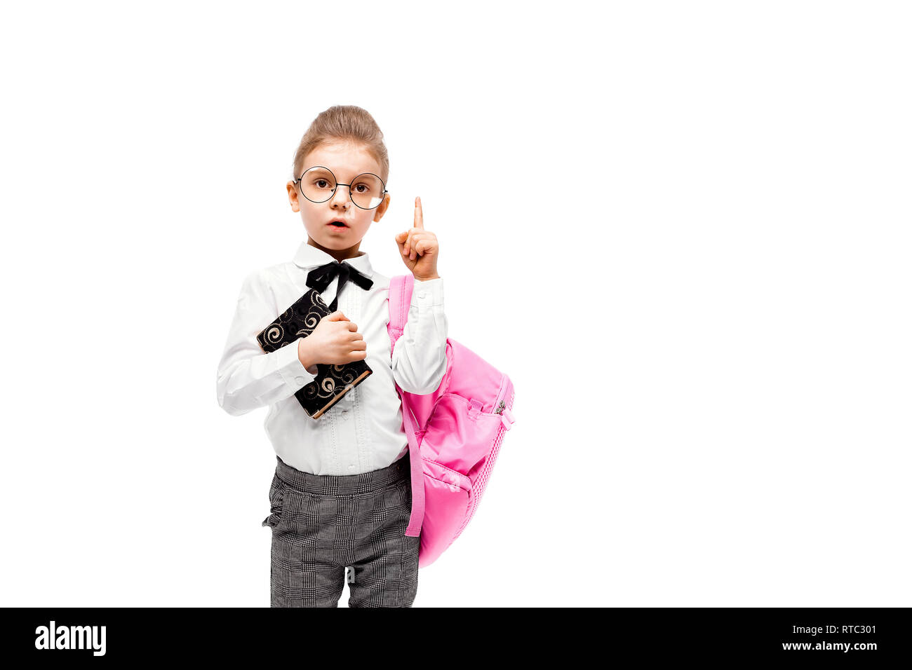 Back to school. Beautiful little girl dressed like a School girl - in white shirt and gray pants, rounded glasses, hold a book, school bag and posing like model points finger up. Isolated on white. Stock Photo