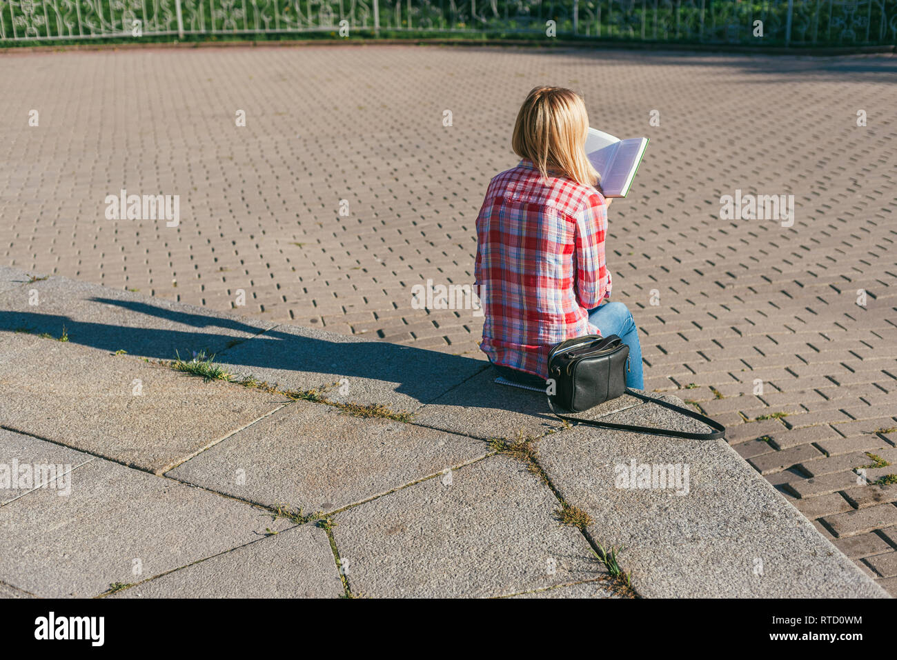 Blond girl in a red checkered shirt is sitting on a stone step and reading a book. Next to her is a black leather handbag. Bright, warm, spring day Stock Photo