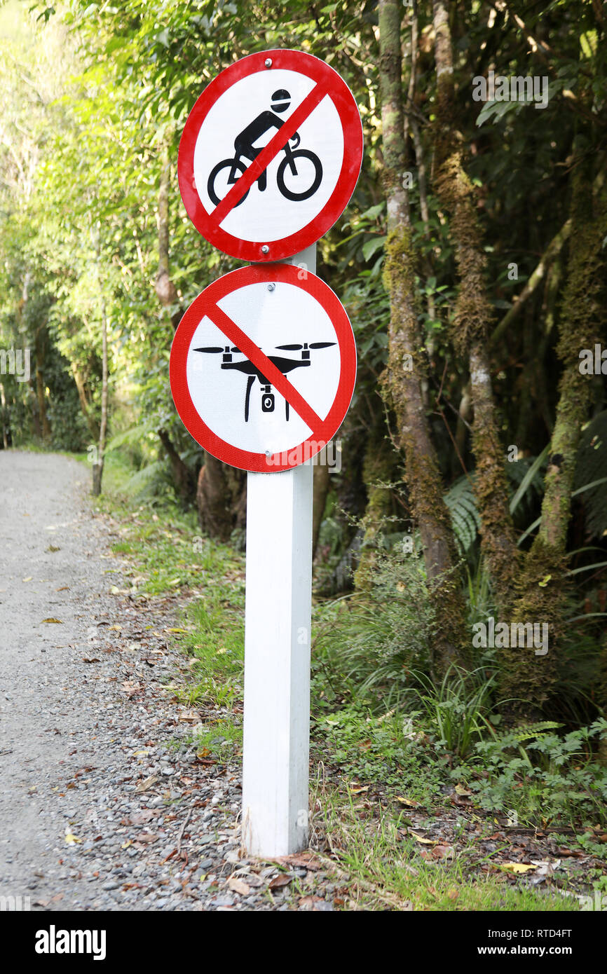 Sign next to the path trail to Franz Josef Glacier saying no drones or cycling with gravel path and trees in background New Zealand South Island Stock Photo