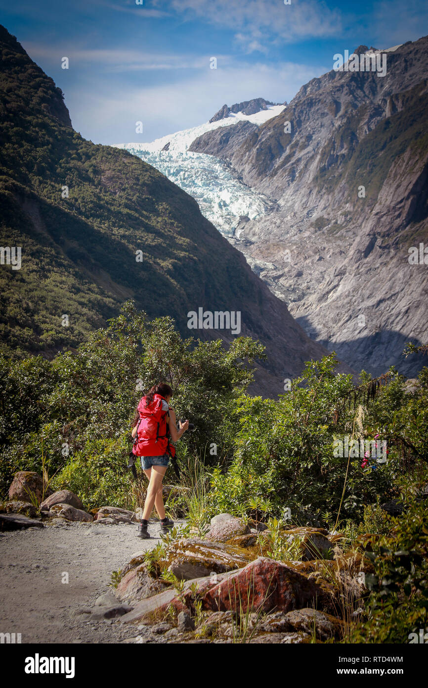 Female tourist hiker walking to Franz Josef Glacier on the path to the glacier with red backpack on a sunny summer day. New Zealand South Island Stock Photo