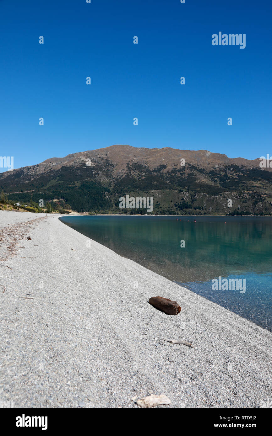 Beautiful Lake Hawea pebble beach with driftwood in summer blue sky clear water pebbles rocks New Zealand South Island Stock Photo