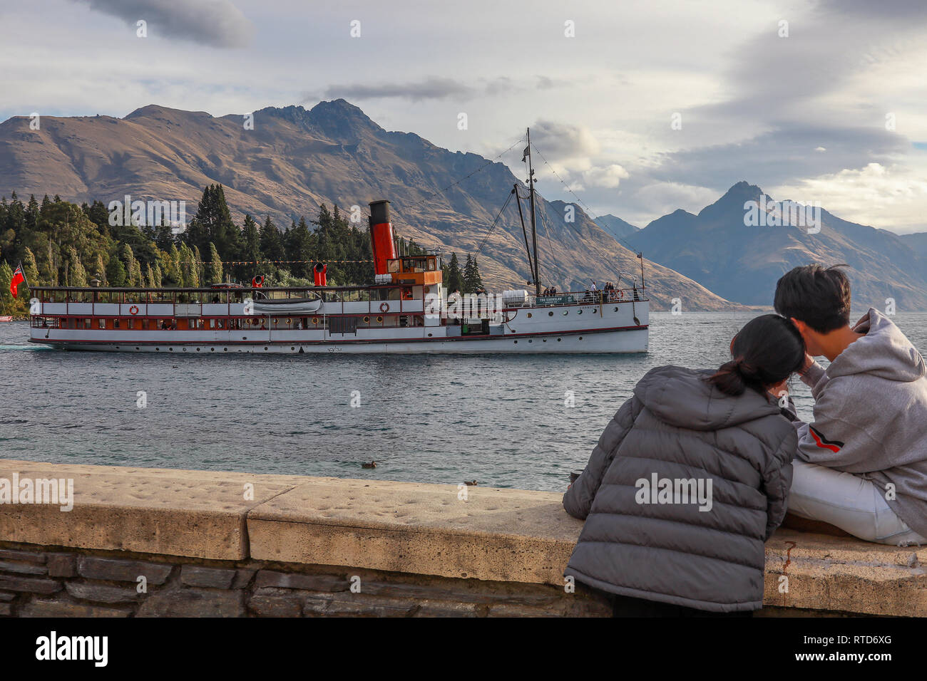 Two Asian tourists watching and filming steam boat Earnslaw on Lake Wakatipu, Queenstown New Zealand South Island with Walter Peak in background Stock Photo