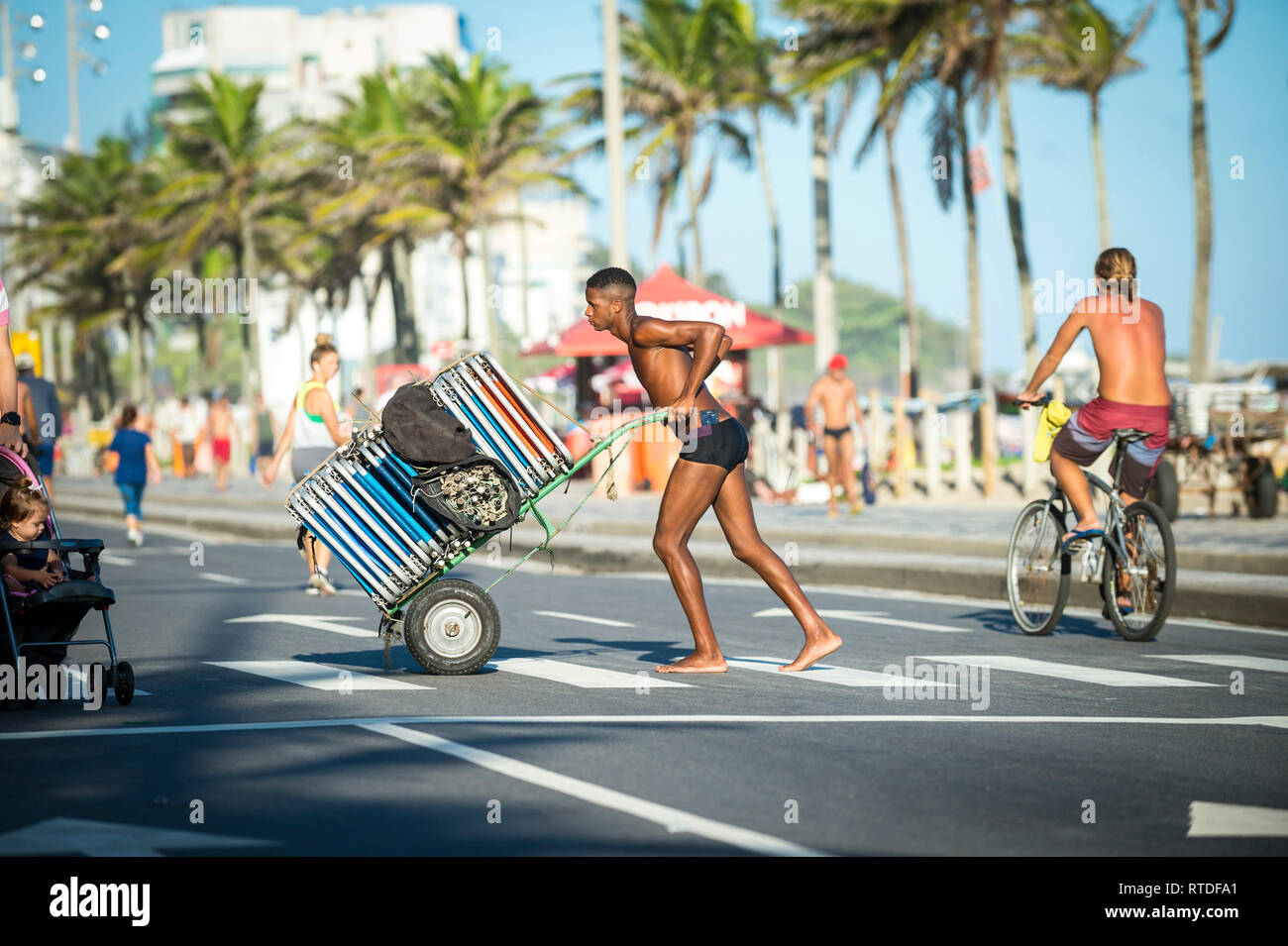 RIO DE JANEIRO - MARCH 26, 2017: A young Brazilian worker pushes a cart of folded chairs from a beach stand across the beachfront road in Ipanema. Stock Photo
