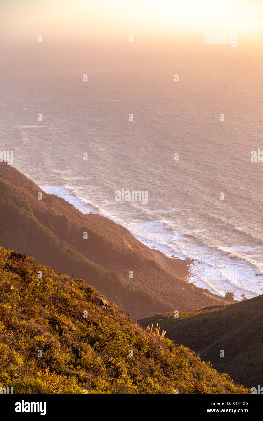 Sunset along the Big Sur coastline in California via a vista view high up in the hills over looking the coast, as waves roll onshore. Stock Photo