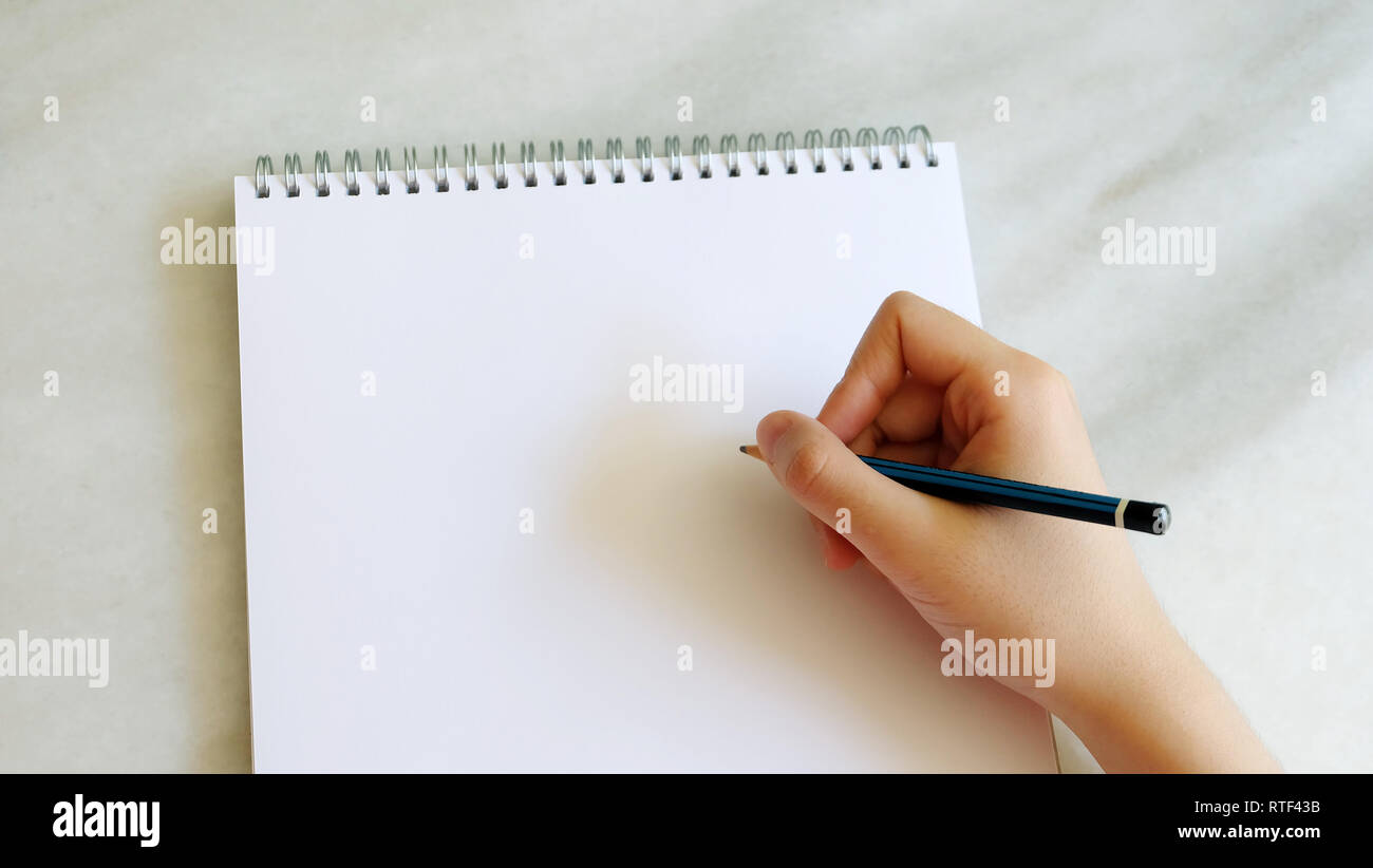 Right hand holding a pencil, on top of a blank sketch book. Stock Photo