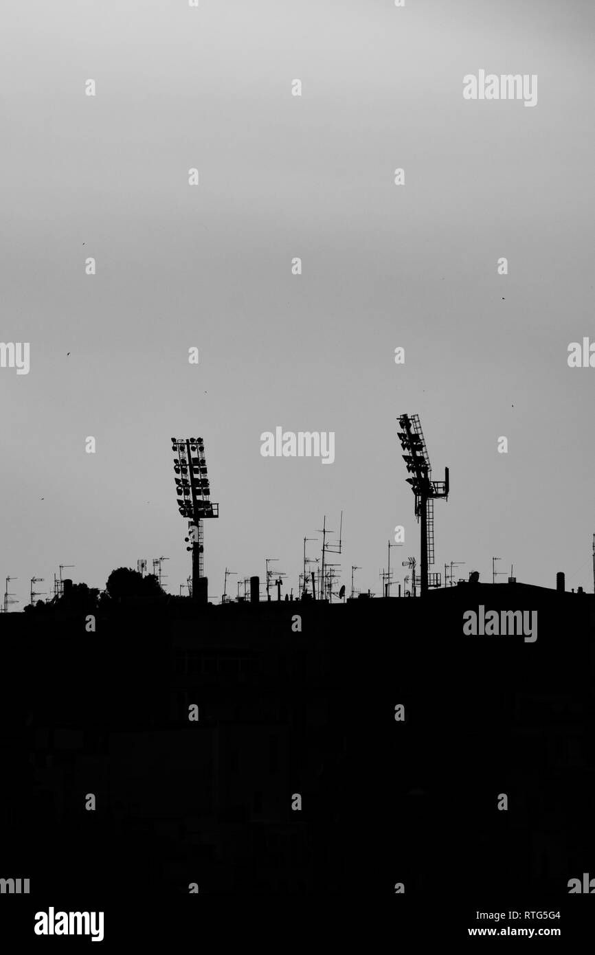 Silhouettes of two sport stadium lights at sunset, town of Matera, Basilicata region, Southern Italy. Black and white picture including some of the city buildings and roofs Stock Photo