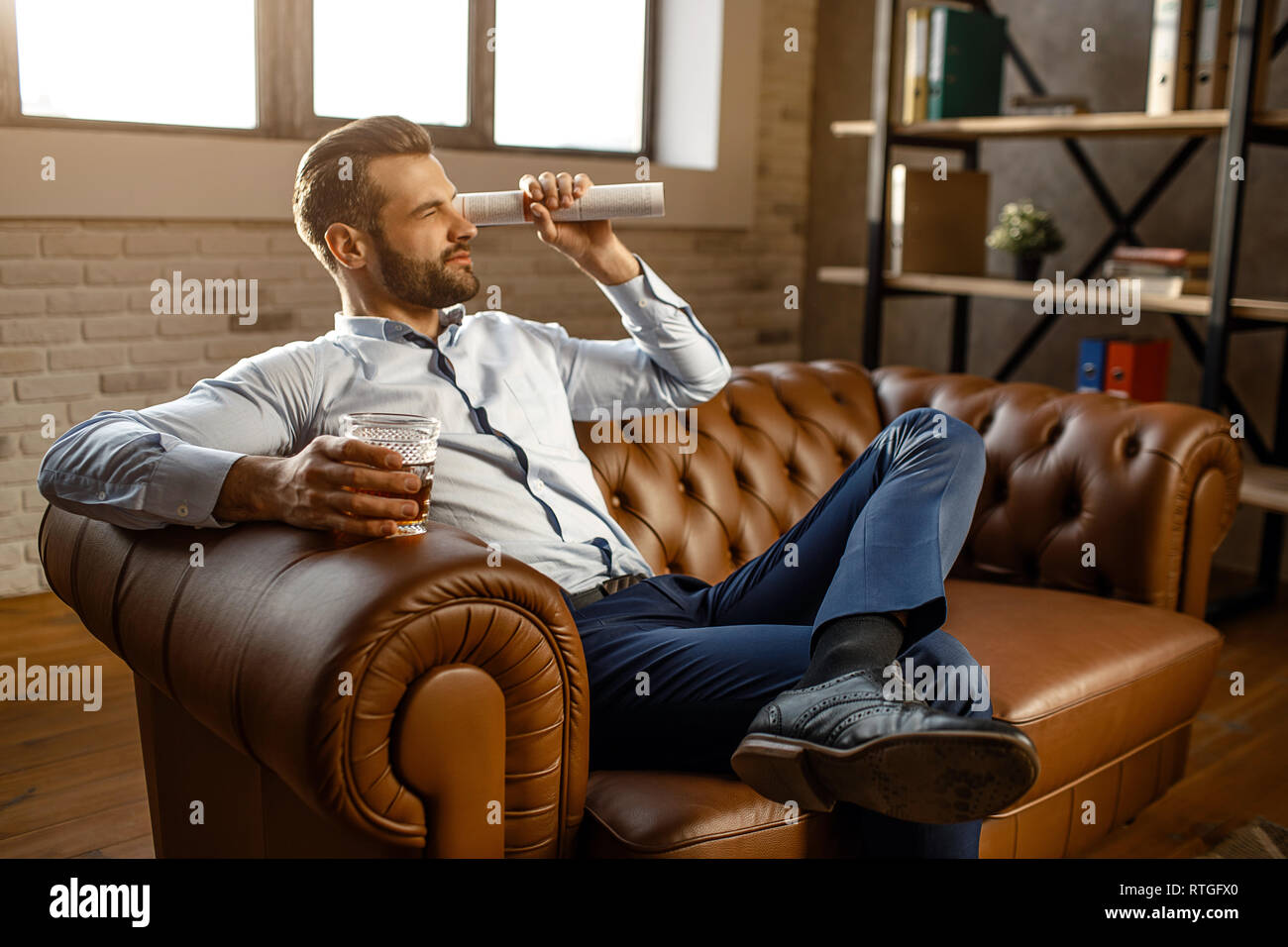 Young handsome businessman sit on sofa and play in his own office. He look  straight through rolled journal. Guy hold glass with whiskey in hand. Leg o  Stock Photo - Alamy