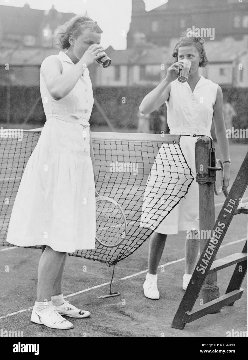 Photo Must Be Credited ©Alpha Press 050000 02/08/1934 Miss D Rowe (Eversley) and Miss M Gardiner (St. Paul's Girls School) after going to 17 deuces in the second set in their Singles march, ordered two orange-ales which they promptly drank. Miss Rowe eventually won 6-3, 4-6, 6-2. The Schoolgirls Lawn Tennis Championships at Queen's Club in West Kensington, London. Stock Photo