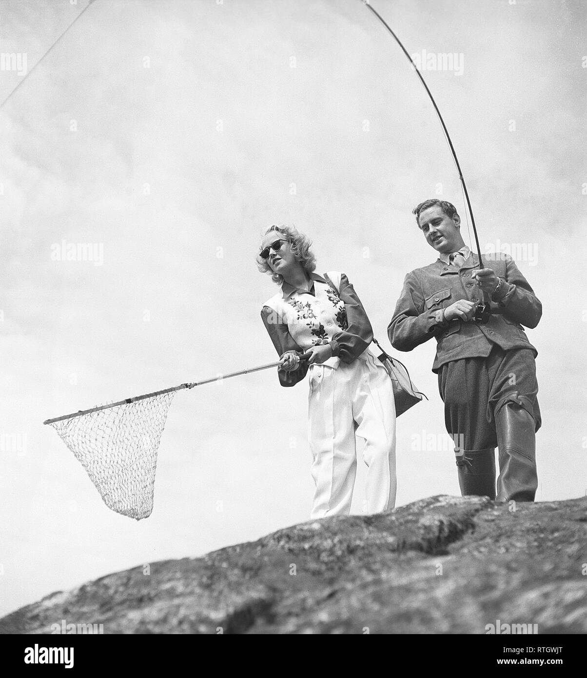 Fishing in the 1950s. A man with a fishing rod looks as if he has a fish on the hook, with his girlfriend ready with a fishnet. Photo Kristoffersson ref BF76-4 Sweden Fishing in the 1950s. A man with a fishing rod looks as if he has a fish on the hook, with his girlfriend ready with a fishnet. Photo Kristoffersson ref BF76-4 Sweden