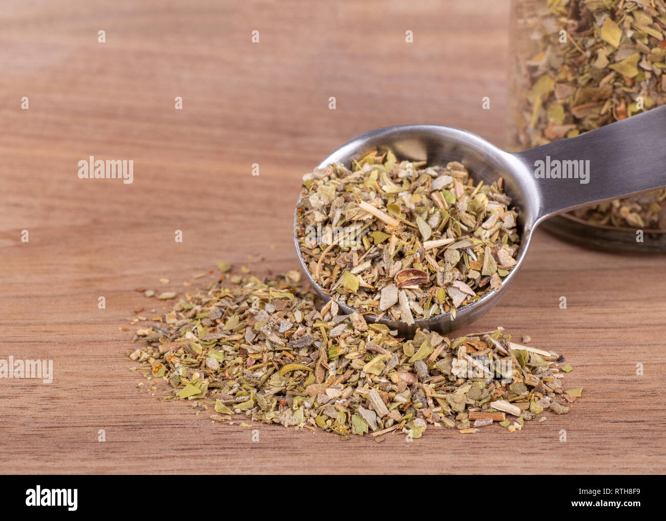 Closeup of a measuring spoon of chopped oregan spilled onto a wooden surface and a jar of herbs in background with copy space Stock Photo