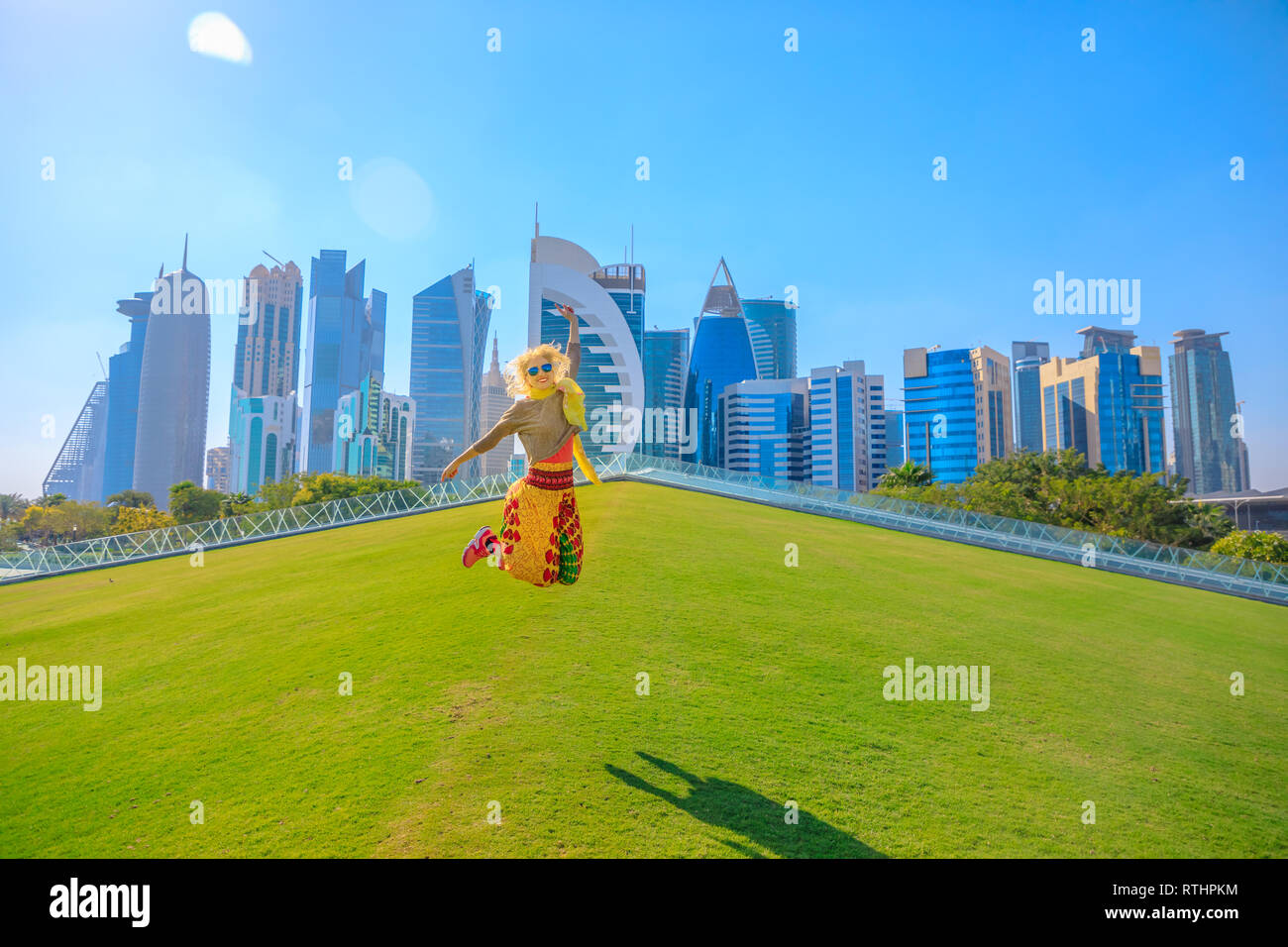 Happy woman traveler on a green lawn in a park jumping with skyscrapers of West Bay on background. Blonde female tourist enjoys Doha Downtown skyline Stock Photo